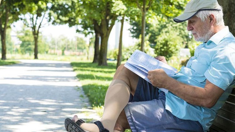 eldre mann iført støttestrømper leser avis på en benk. foto. 
